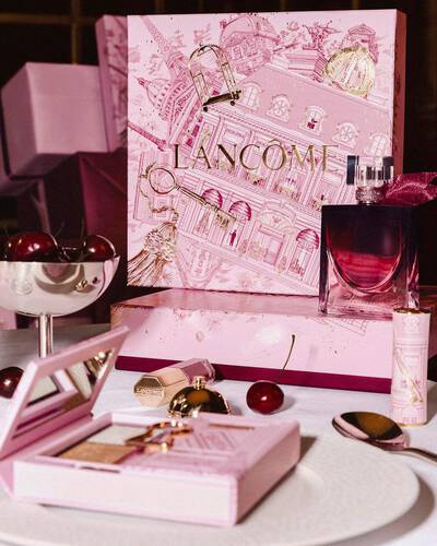 Świąteczna kolekcja Lancome wyeksponowana na stole, z dużym, rożowym pudełkiem prezentowym, perfumami, kosmetykami do makijażu i świeżymi wiśniami.