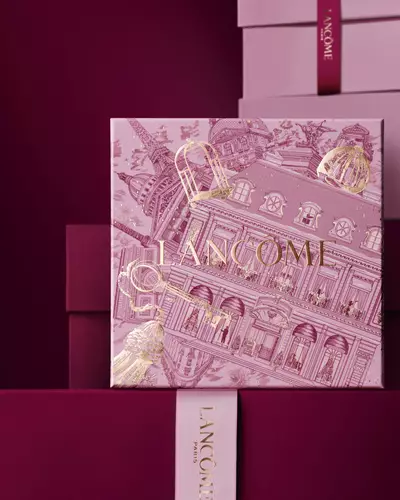 Zdjęcie rożowego świątecznego pudełka z Lancome stojącego przed stosem rożowych i ciemnoczerwonych pudełek, na ciemnoczerwonym tle.