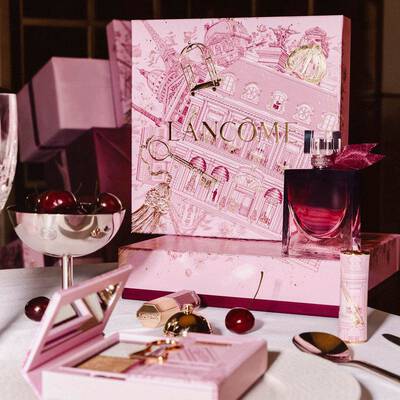 Świąteczna kolekcja Lancome wyeksponowana na stole, z dużym, rożowym pudełkiem prezentowym, perfumami, kosmetykami do makijażu i świeżymi wiśniami.