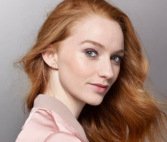 A close-up portrait of a young woman with long, wavy red hair, fair skin, and blue eyes, smiling softly while wearing a light pink jacket.