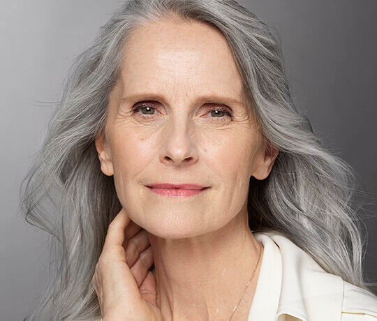 A close-up portrait of an older woman with long, wavy gray hair and a gentle smile, looking directly at the camera. She has a natural makeup look and is dressed in a light-colored top.