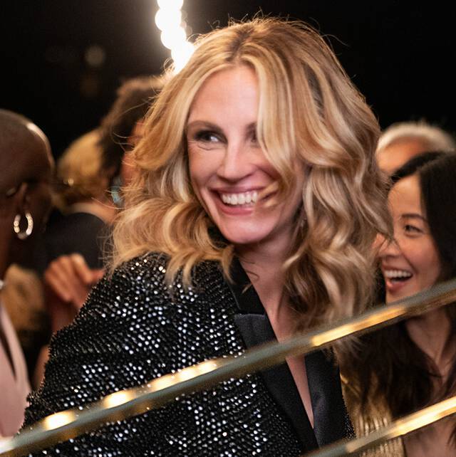 Uśmiechnięta kobieta z blond włosami w eleganckiej kreacji, otoczona ludźmi, w radosnej atmosferze.