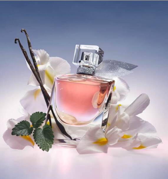 Szklana butelka perfum w rożowym kolorze otoczona białymi kwiatami i gałązką wanilli.