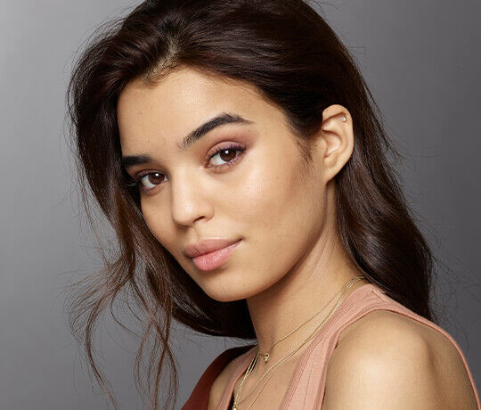 A young woman with long, wavy brown hair and natural makeup is looking over her shoulder, giving a soft smile. She is wearing layered gold necklaces and has an elegant appearance. The background is neutral and softly lit.