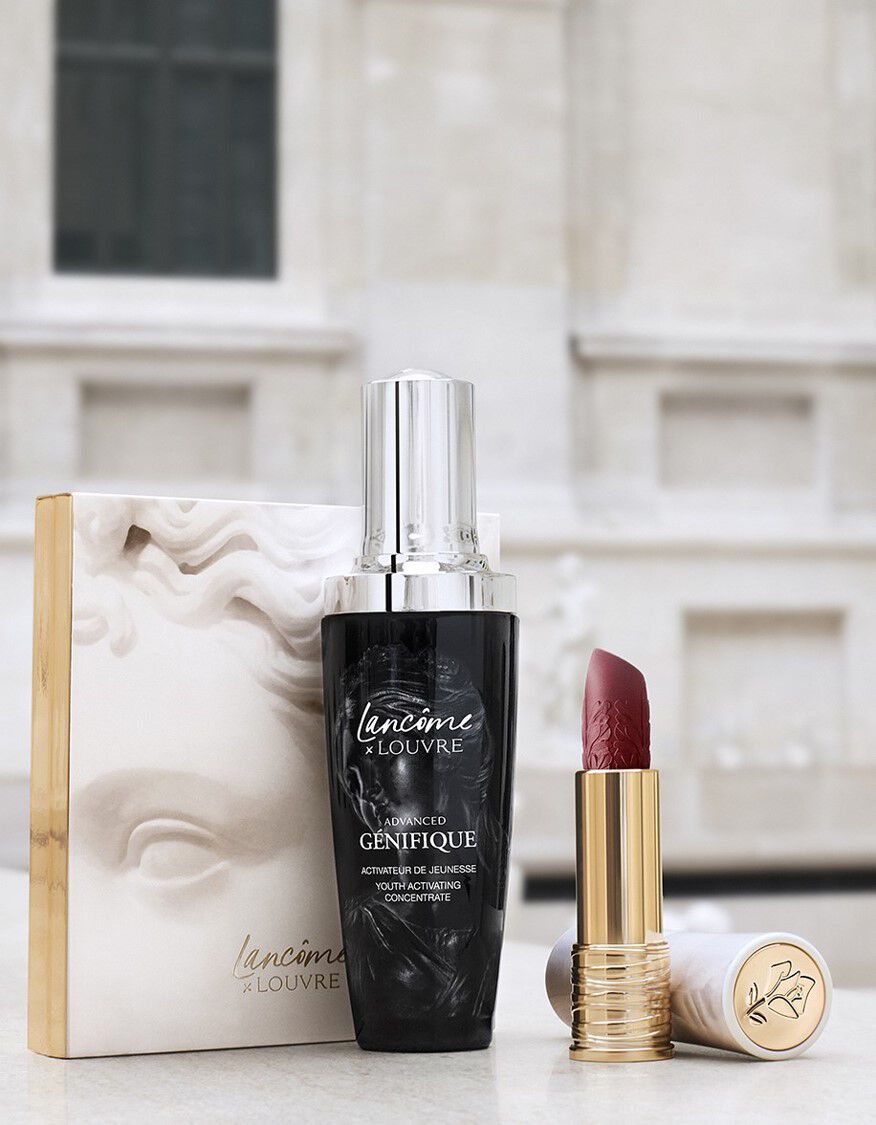 A bottle of Lancome advanced serum and a red lipstick sit next to a decorative box featuring a sculpture, with a blurred artistic background resembling the Louvre.
