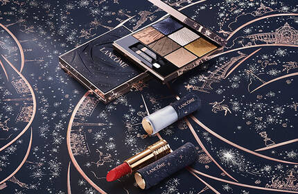Alt text: A flat lay featuring makeup products, including eyeshadow, lipstick, and nail polish, arranged on a dark background with whimsical white starburst and spider illustrations.