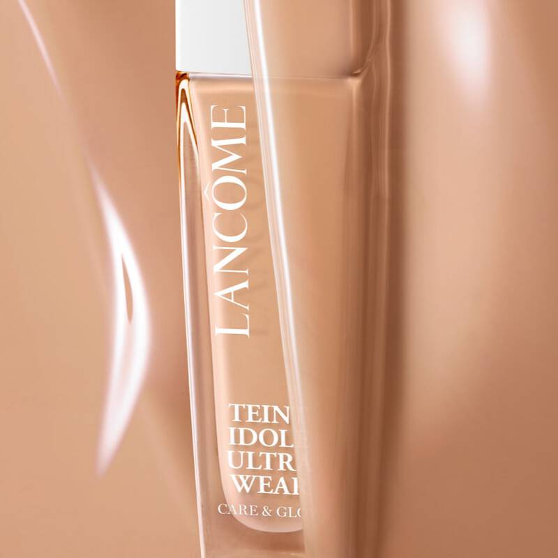 A close-up of a Lancome foundation bottle partially obscured in a warm-toned background. The bottle is sleek and elegant, labeled "Teint Idole Ultra Wear."