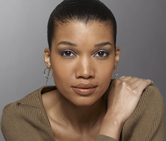 A close-up portrait of a woman with dark hair pulled back, wearing gold hoop earrings and a neutral-toned top. She has a natural makeup look, with soft eye makeup and a nude lip, and is gazing directly at the camera.
