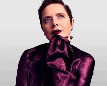 A woman with short hair poses thoughtfully, wearing a deep purple blouse. She has a dark lip color and gazes upward, resting her chin on her hand. The background is a soft gradient.