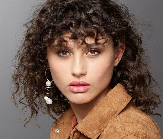 A close-up portrait of a person with curly hair, wearing large, pearl-accented earrings and a suede jacket. The background is a neutral gray. The subject has a natural makeup look, featuring a subtle lip color.