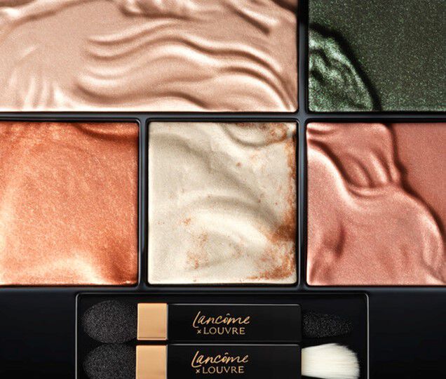 A close-up view of a makeup palette featuring various shimmering eyeshadow shades in warm and cool tones, arranged in distinct sections. Two double-ended applicators are included at the bottom.