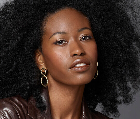 Close-up portrait of a young woman with natural, voluminous curls, wearing gold hoop earrings and a leather jacket, looking directly at the camera with a soft expression.