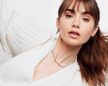 A young woman with long, brown hair styled in a ponytail is wearing a white, pleated top and a delicate necklace. She gazes directly at the camera with a soft expression.