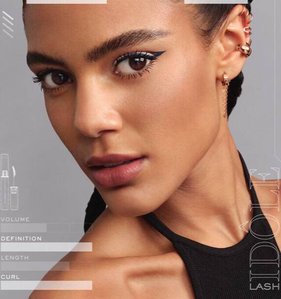 A close-up portrait of a woman with stylish makeup, featuring bold eyeliner and defined eyelashes. She has braided hair, a strong jawline, and is wearing a black sleeveless top. The background is neutral, enhancing her features.