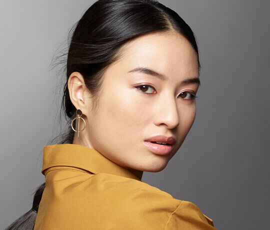 A close-up portrait of a woman with long, dark hair wearing a mustard-colored shirt and circular earrings, looking over her shoulder with a subtle expression.