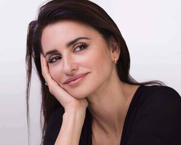 A woman with long, dark hair rests her chin on her hand, smiling gently with a thoughtful expression. She is wearing a black top and has subtle makeup, with a soft background.
