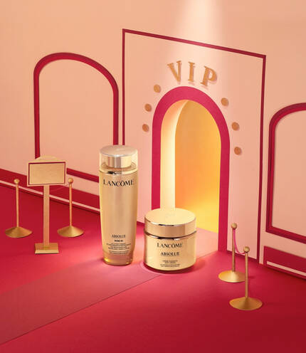 A gold and pink display featuring Lancome's Absolue skincare products, including a serum bottle and a cream jar, alongside a small stand and a decorative frame. The background has a geometric design.
