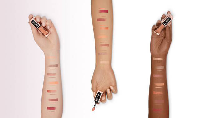 Three arms with swatches of various lipstick shades on their skin. The first arm on the left shows lighter shades, the middle arm holds a lipstick while displaying medium shades, and the third arm on the right features darker shades.