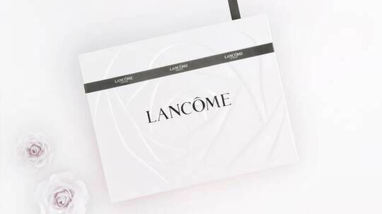 Biała pudełko kosmetykow Lancome z logo, z delikatnymi rożami w tle.