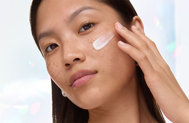 A person with freckles applying cream on their cheek, looking confidently at the camera.