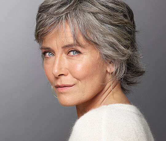 A close-up portrait of a woman with short, silver hair and blue eyes, wearing a soft white sweater. She is looking directly at the camera with a slight smile.