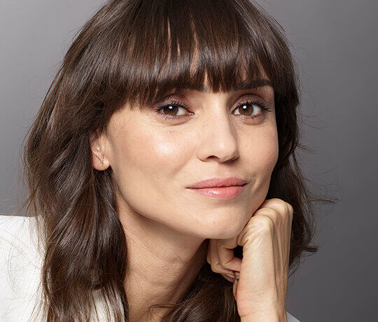 A close-up portrait of a woman with long, dark hair and a subtle smile, resting her chin on her hand. She has a smooth complexion and a natural makeup look, with soft lighting highlighting her features.
