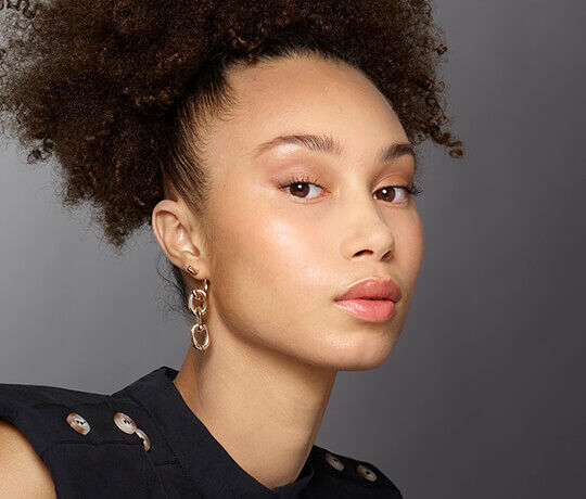 A close-up portrait of a person with curly hair styled in a high bun, wearing gold earrings and light makeup, looking directly at the camera.