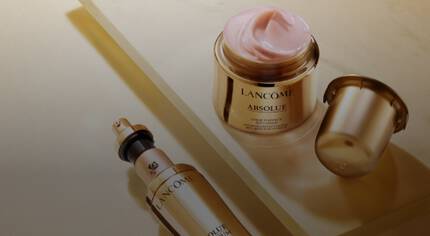 Zestaw kosmetykow Lancome Absolue, w tym serum w szklanej butelce z pompką i rożowym kremem w eleganckim słoiczku, na tle złotego powierzchni.