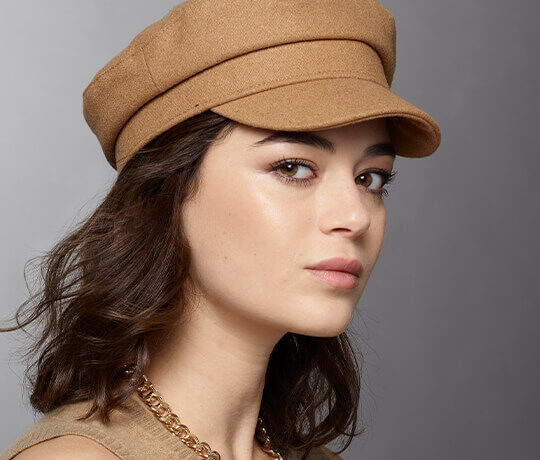 A profile view of a young woman wearing a tan hat and a gold chain necklace, with soft makeup and wavy hair.