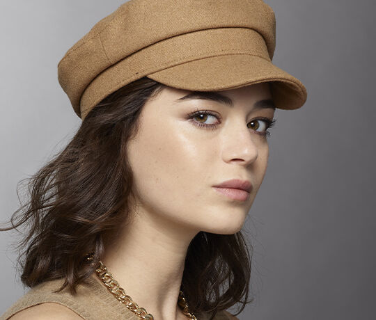 A portrait of a young woman wearing a brown cap, gazing slightly to the side. She has a soft complexion, defined features, and is accessorized with a chain necklace.