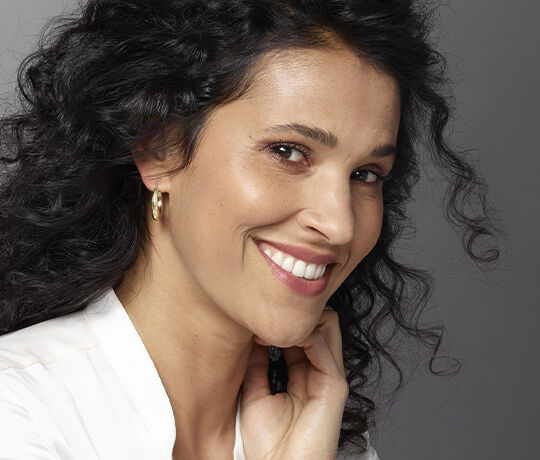 A smiling woman with curly black hair, wearing hoop earrings, poses gracefully against a neutral background.