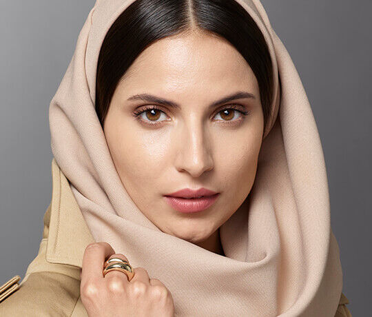 A close-up portrait of a woman with a soft expression, wearing a beige hijab and a simple ring on her finger. The background is muted and neutral, highlighting her features.