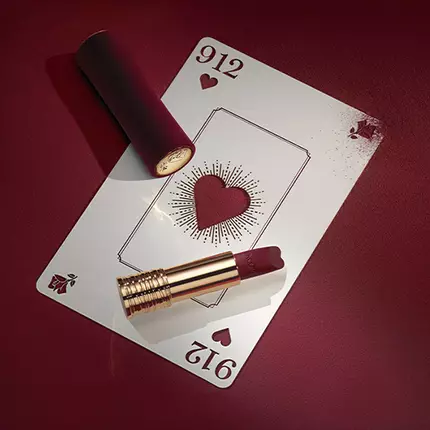 A playing card with a heart design lies on a deep red surface, next to a tube of lipstick in a matching shade. The card features ornate details, enhancing the elegant aesthetic.