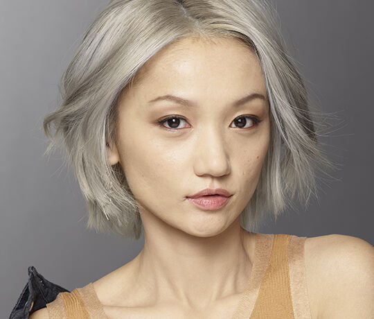 A close-up portrait of a person with short, silver hair and a natural makeup look, featuring a soft expression and clear skin. The background is neutral and softly lit.