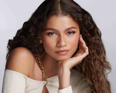 A young woman with long, wavy hair is posed thoughtfully, resting her chin on her hand, wearing a stylish off-the-shoulder top. Her expression is confident and serene against a softly blurred background.
