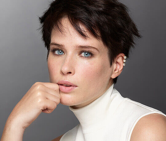 A close-up portrait of a woman with short hair and striking blue eyes, wearing a high-necked, sleeveless top. She rests her chin on her hand, showcasing a subtle makeup look.