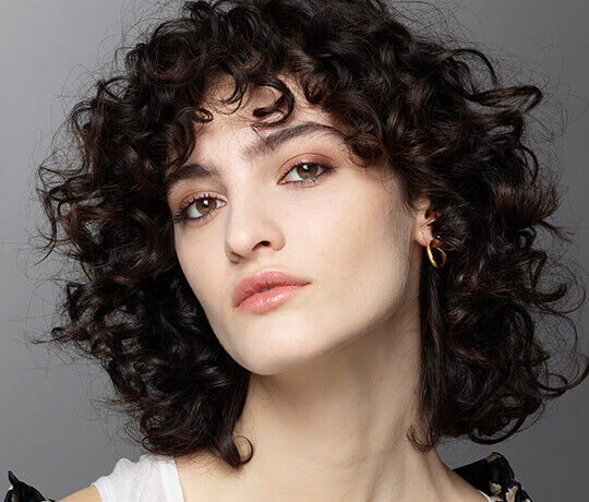 Close-up of a model with curly hair and a subtle makeup look, wearing gold earrings and a light-colored top.