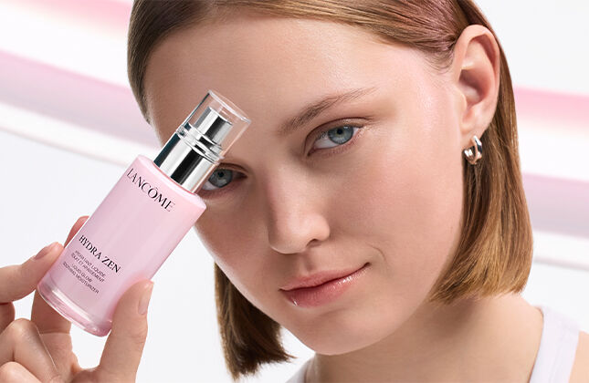 A woman with short hair holds a pink skincare bottle near her face, showcasing a fresh and elegant beauty look against a soft, flowing background.