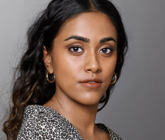 A close-up portrait of a young woman with long, dark hair, wearing hoop earrings and a patterned top. She has a neutral expression and natural makeup, against a grey background.