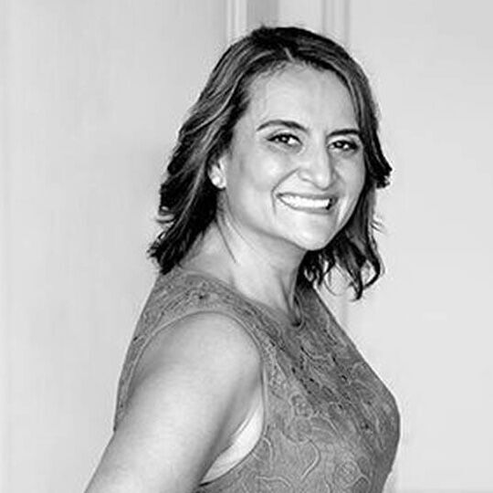 A smiling woman with shoulder-length hair, wearing a sleeveless dress, poses against a light background.