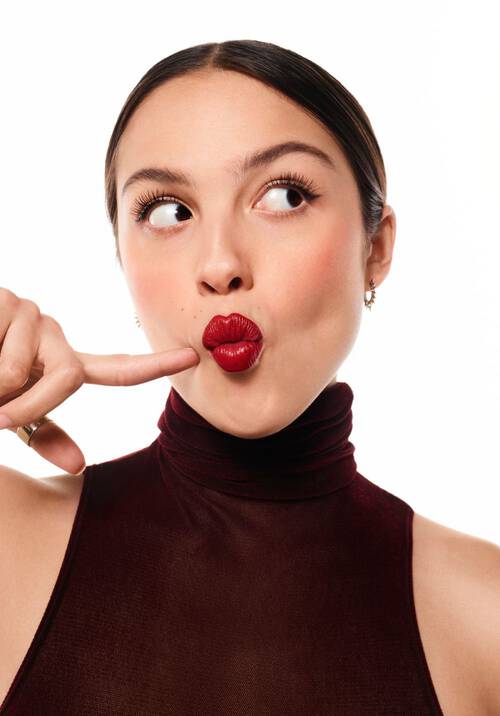 A young woman with long hair styled in a sleek bun, wearing a maroon turtleneck, playfully poses with a finger to her lips, showcasing bold red lipstick and a whimsical expression.