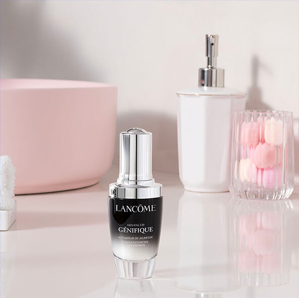 Butelka serum z logo Lancome, umieszczona na jasnej powierzchni obok kwiatowej dekoracji w wazonie i innych kosmetykow.