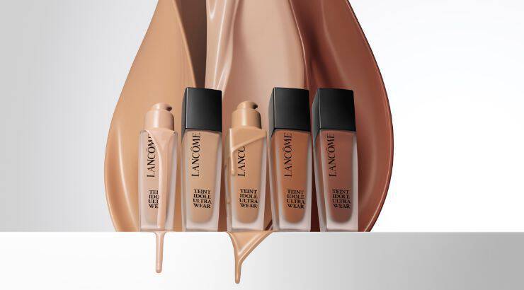 A collection of Lancome foundation bottles displayed in front of flowing liquid foundation swatches in various shades, showcasing the brand's Tient Idole Ultra Wear range.