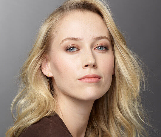 A close-up portrait of a woman with long, wavy blonde hair and blue eyes, wearing subtle makeup and delicate earrings, gazing directly at the camera.