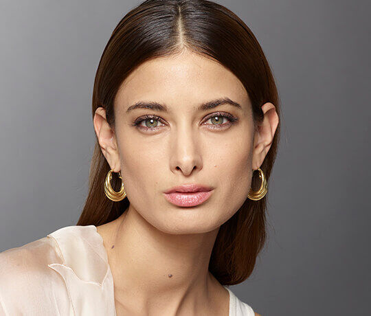 A close-up portrait of a model with long hair, wearing large gold hoop earrings and a natural makeup look, gazing directly at the camera.