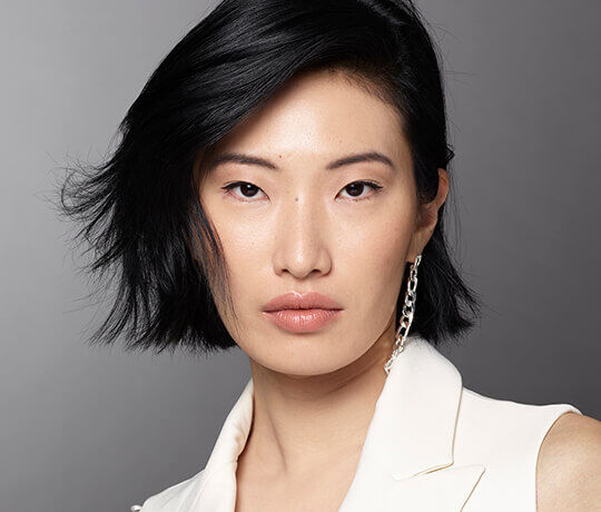 A close-up portrait of a model with short black hair, wearing a white blazer. She has a neutral expression and subtle makeup, showcasing her natural features. A statement earring is visible on one side.