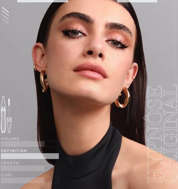 A close-up image of a woman showcasing flawless makeup, with emphasis on her defined eyelashes and smooth skin. She has dark, straight hair and is wearing bold earrings. The text on the side highlights features related to mascara, such as volume and definition.