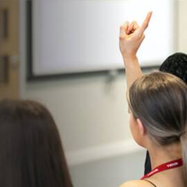 A classroom scene showing students from behind, with one student raising their hand to ask a question.