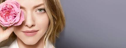 A woman holds a pink rose to her eye, smiling softly against a gray background. Her hair is styled in loose waves, and she has a natural makeup look.