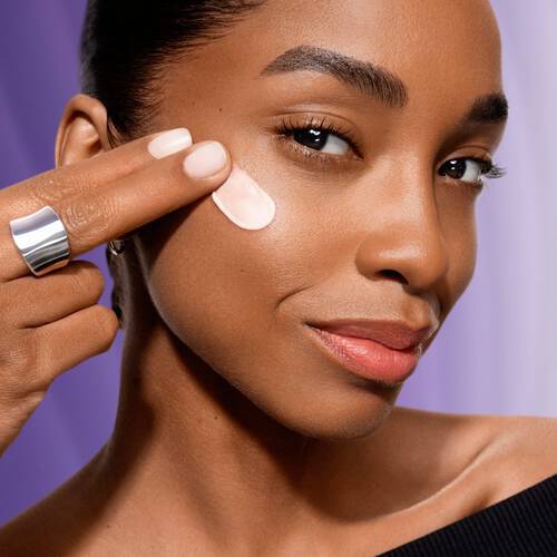 Kobieta nakłada krem na twarz, uśmiechając się, z delikatnym makijażem, w tle fioletowe światło.